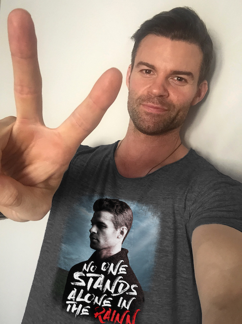 Television actor Daniel Gillies portrait. He is wearing a special edition RAINN t-shirt. The t-shirt features Daniel's face and the words "No one stands alone in the RAINN." He holds up a peace sign to the camera. 